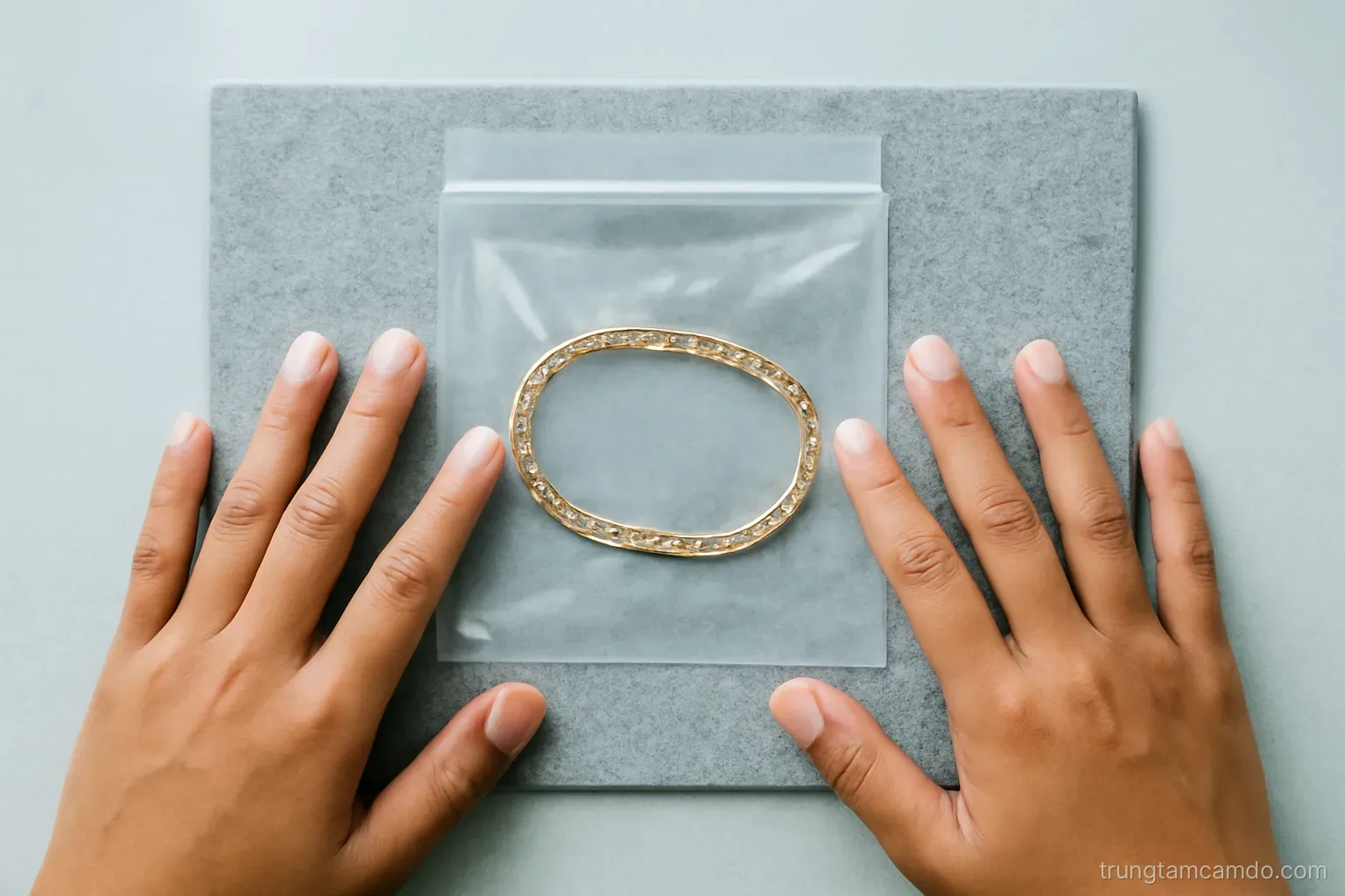 Top-down shot of a clear sealed pouch holding a gold bracelet on a grey felt mat, two Vietnamese hands pressing thumbprints beside it, cool daylight, muted grey and gold palette